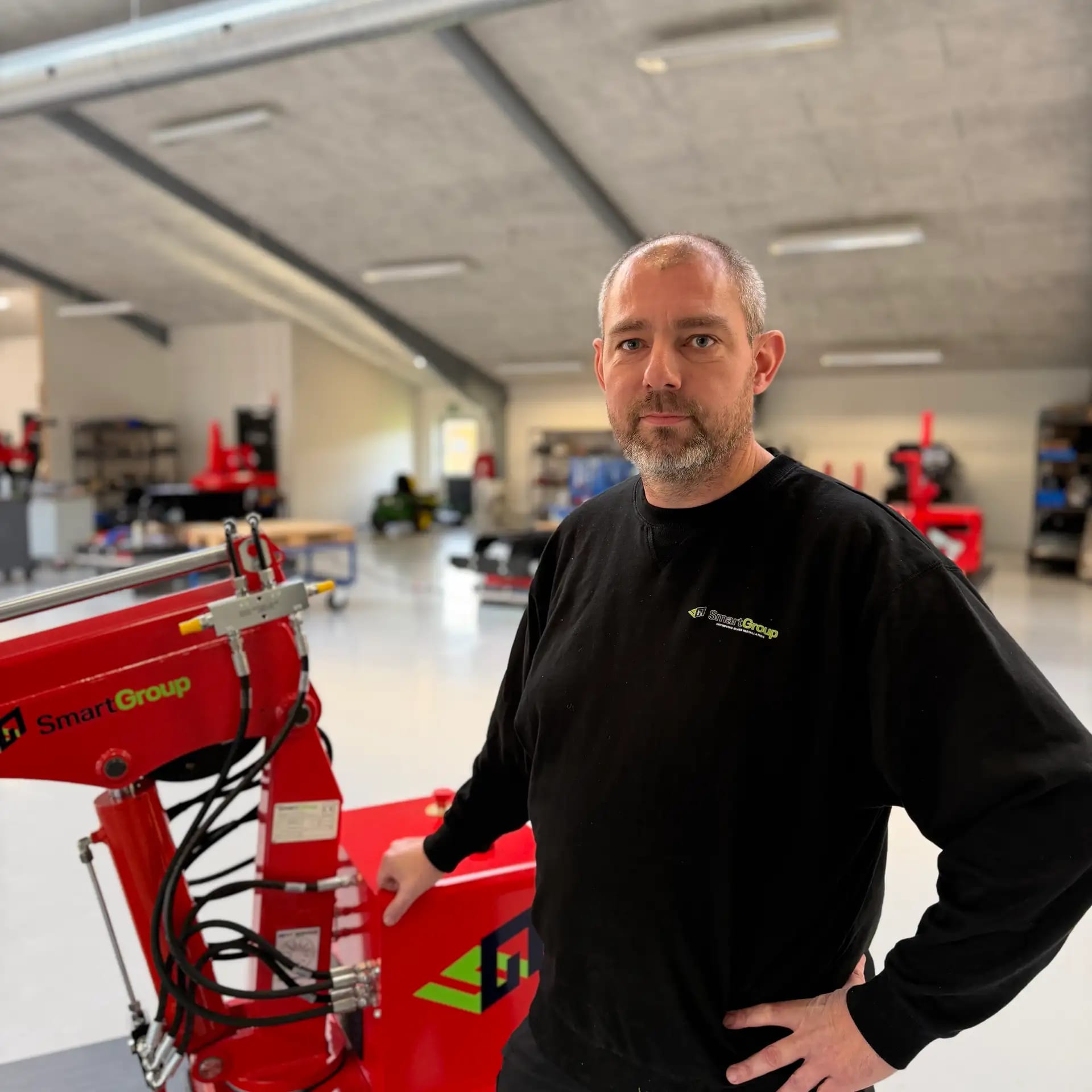 En mand med skæg står indendørs ved siden af rødt industrielt udstyr iført en sort sweater med logo. Baggrunden viser et rummeligt, moderne værksted med hylder, borde og yderligere maskiner.